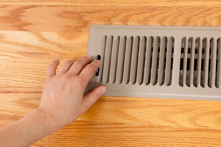 Horizontal photo of female hand opening up heater floor vent with Red Oak Floors in background.